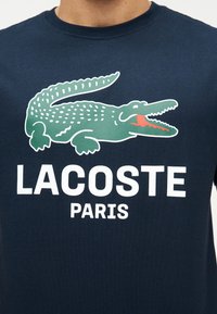Πράσινο λογότυπο κροκόδειλου με ανοικτό στόμα πάνω από το λευκό κείμενο «LACOSTE PARIS» σε ύφασμα ναυτικού μπλε χρώματος.