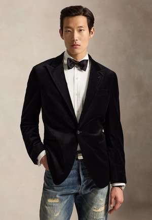 Ung mand iført en sort fløjlsblazer, hvid skjorte, sort butterfly og slidte blå jeans står foran en neutral baggrund.