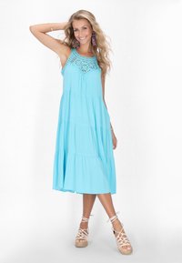 Rochie bleu deschis fără mâneci, cu detalii din dantelă la bust, design în straturi fluide, asortată cu sandale cu talpă înaltă din bej.