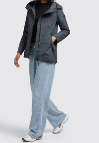 Wasserabweisende graue Outdoor-Jacke mit Kapuze, Reißverschluss vorne und seitlichen Taschen. Kombiniert mit hellblauen weiten Jeans und weißen Turnschuhen.