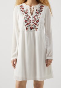 Vit långärmad klänning med blommig broderi i rött och rosa. Har en knytkrage och en flödande silhuett med en texturerad tyg.