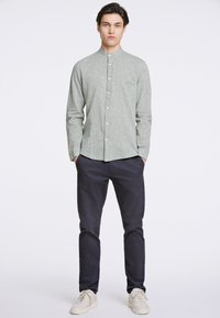 Camicia verde a maniche lunghe con texture, colletto button-down, taschino singolo sul petto, abbinata a pantaloni blu navy e sneakers chiare.