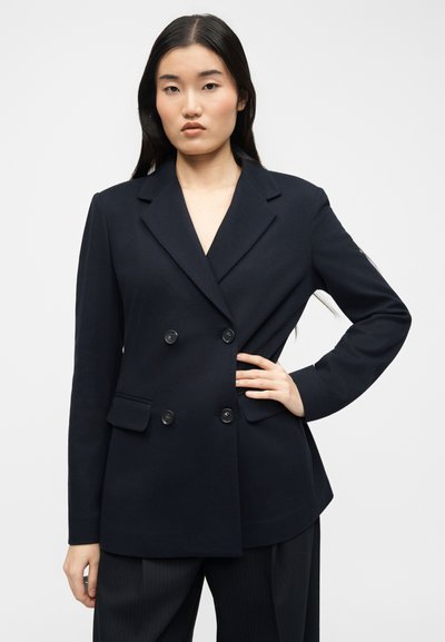 Γυναίκα που φοράει διπλόμπουτο μαύρο σακάκι με τέσσερα κουμπιά, στέκεται με το ένα χέρι στη μέση της μπροστά σε έναν απλό φόντο.