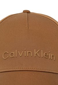 Casquette marron Calvin Klein en tissu texturé, avec un logo en relief à l'avant et une visière cousue. Dessus arrondi avec un détail de bouton.