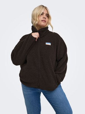 Femme aux cheveux blonds portant une veste polaire marron foncé et un jean bleu, tenant la fermeture éclair de la veste près du menton avec une main.
