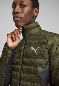 Grønne puffede jakke med høj krave, med lynlås lukning, sorte detaljer på ærmerne og et reflekterende Puma-logo. Tekstureret overflade.