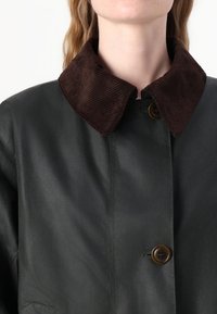 Casaco verde escuro com gola em bombazine castanha texturada, fecho de um único botão e acabamento liso e mate.