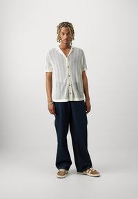 Camisa blanca de manga corta y punto abierto con botones, combinada con jeans de pierna ancha azul oscuro y zapatillas marrones con rayas blancas.