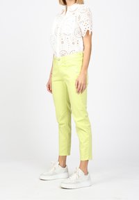 Vit broderad blus med korta ärmar, kombinerad med ljusgula jeans. Vita sneakers med texturerade detaljer. Modellen står i profil.