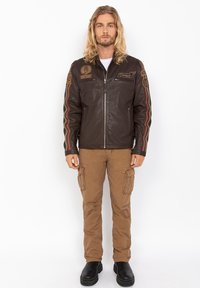 Giacca in pelle marrone con chiusura a zip, strisce laterali e diverse toppe. Abbinata a pantaloni cargo beige e stivali neri.