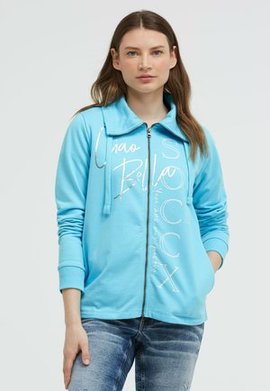 Frau mit langen braunen Haaren, die einen hellblauen Kapuzenpullover mit weißem Schriftzug und ausgewaschene blaue Jeans trägt, steht mit einer Hand in der Tasche.