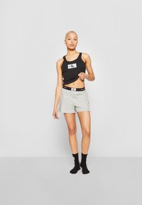 Persona joven con cabello corto rubio viste una camiseta sin mangas negra, pantalones cortos grises y calcetines negros, frente a un fondo gris liso.