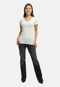 Hvid tætsiddende t-shirt med "Guess" i lyseblå skrifttype, parret med mørke lige ben jeans og sorte sko, på en model foran en ensfarvet baggrund.