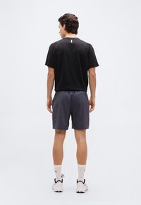 Άνδρ ας που στέκεται με την πλάτη, φοράει μαύρο μπλουζάκι, γκρι σορτς, λευκές κάλτσες και λευκά αθλητικά παπούτσια σε απλό λευκό φόντο.