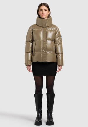 Nafouklá olivově zelená bunda s vysokým límcem, zapínáním na zip a bočními kapsami, kombinovaná s černou žebrovanou sukní a černými kozačkami až ke kolenům.