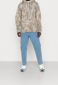 Beige camo pullover hoodie med snoretræk, elastiske manchetter, parret med lyseblå denimbukser og grå-hvide træningssko.