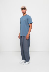 Homme portant une casquette beige, un t-shirt bleu à manches courtes, un pantalon gris ample et des baskets blanches se tenant devant un fond blanc uni.
