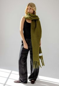 Écharpe vert olive en matériau doux, avec des bords à franges et une étiquette. Associée à un pantalon large noir à motifs et des chaussures foncées.