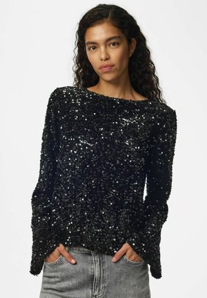 Femme aux cheveux bouclés portant un haut noir à manches longues recouvert de sequins brillants et un jean gris, debout les mains dans les poches.