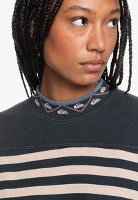 Dark sweater with horizontal cream stripes, textured collar featuring a patterned detail of triangles, and layered blue shirt underneath.