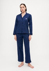 Marineblå pyjamassett laget av glatt stoff, med en knapp-up topp med lomme og rullede mansjetter på buksene. Vertikale striper tekstur.