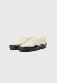 Canvas sneakers in off-white with black rubber soles. Features white laces and a blue logo tag on the side. Rounded toe design.