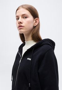 Νεαρή γυναίκα με ίσια ανοιχτό καστανά μαλλιά, που φορά ασημένια σκουλαρίκια κρίκους και μαύρο φούτερ με φερμουάρ με το λογότυπο "BOSS".