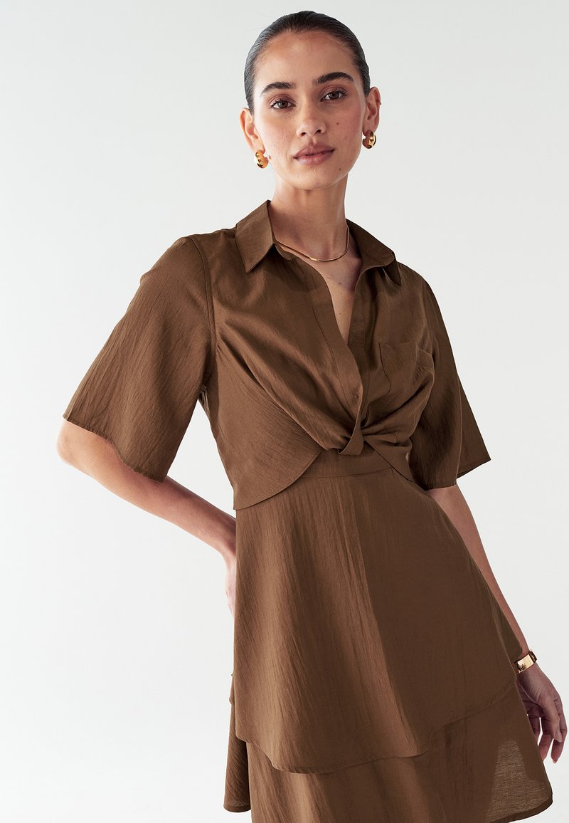 Robe brune à manches courtes avec un design à l'avant torsadé et un col. Fabriquée en tissu léger avec une texture lisse, elle présente des détails de jupe superposée.
