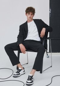 Černá sako přes bílé tričko s textem, kombinované s černými kalhotami a černo-bílými teniskami. Hladká textura, moderní design.