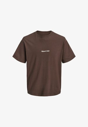 Uśmiechnięte dziecko stoi ubrane w kraciastą koszulę na brązowym t-shircie, szerokie niebieskie dżinsy i czarne buty na jednolitym jasnym tle.