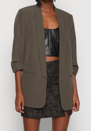 Femme portant un blazer ample vert olive sur un crop top en cuir noir et une mini jupe noire texturée à motifs floraux.