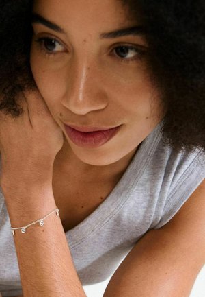 Silbernes Armband mit anhängenden Charms, das eine zarte Kette und kleine dekorative Elemente aufweist. Getragen an einem gebräunten Handgelenk gegen ein graues Oberteil.