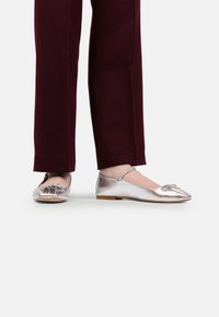 Balerini metalici argintii cu detaliu în formă de fundă, talpă plată și vârf rotunjit, asortați cu pantaloni burgundy pe un fundal deschis.