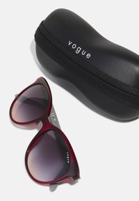 Raudonais atspalviu pagaminti dideli akiniai nuo saulės su gradientinėmis lęšiais, kartu su juodu užtrauktuku uždaromu apsauginiu dėklu, ant kurio yra "vogue" logotipas.