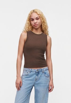 Jeune femme aux longs cheveux blonds bouclés, portant un haut marron sans manches et un jean bleu clair, debout devant un fond blanc uni.