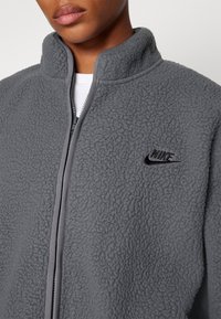 Giacca in pile grigia con colletto alto, tessuto testurizzato, chiusura a cerniera completa e un piccolo logo Nike nero sul petto.