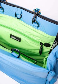 Lichtblauwe tasinterieur met een levendige groene voering, een ritscompartiment, zwarte hardware en een gestructureerde stof. Sluiting met kleurrijke accenten.