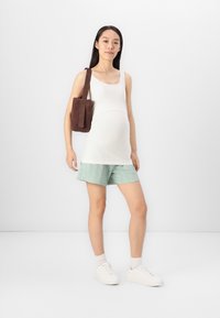 Μαμά φανέλα σε λευκό βαμβάκι, συνδυασμένη με μίντ-πράσινα σορτσάκια με υφή. Το μοντέλο κρατά μια καφέ σουέτ τσάντα ώμου και φοράει λευκά αθλητικά παπούτσια.