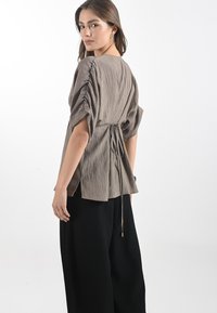 Top marrone chiaro con texture, dal fit rilassato, maniche a sbuffo e laccio regolabile in vita. Abbinato a pantaloni neri larghi.