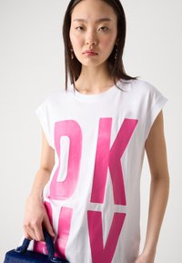 T-shirt bianco in cotone con vestibilità oversized, caratterizzato da una grande grafica rosa "DKNY". Il modello tiene una piccola borsa blu con finitura testurizzata.