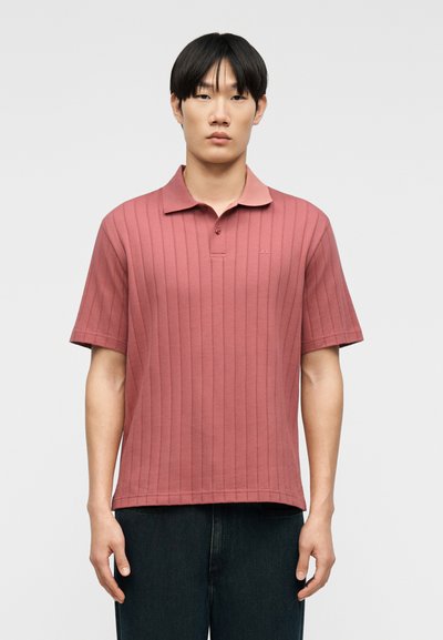 Jeune homme portant un polo côtelé rose à manches courtes et un pantalon foncé, debout devant un fond clair uni.
