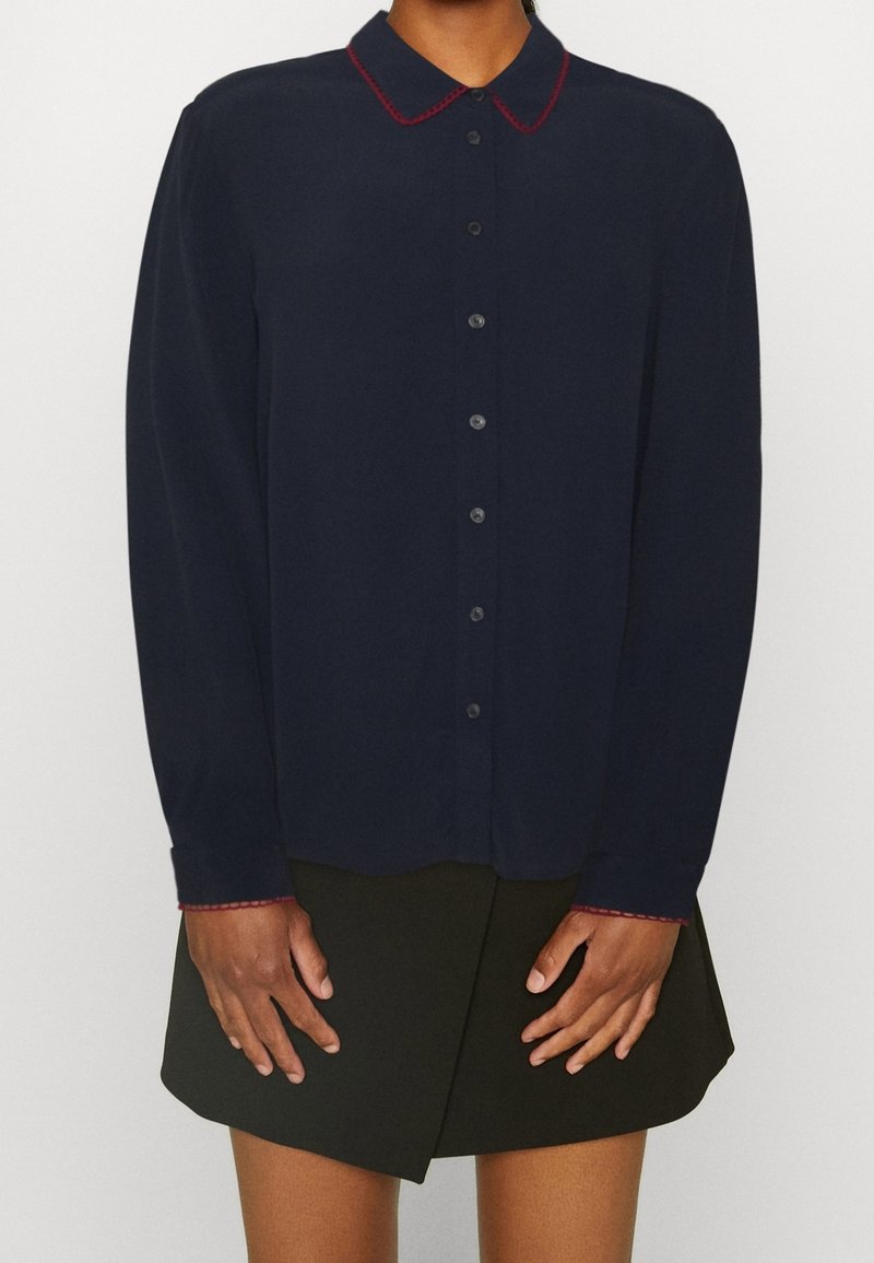 Personne portant une chemise bleu marine à boutons avec des coutures rouges sur le col et les poignets, associée à une mini-jupe noire.