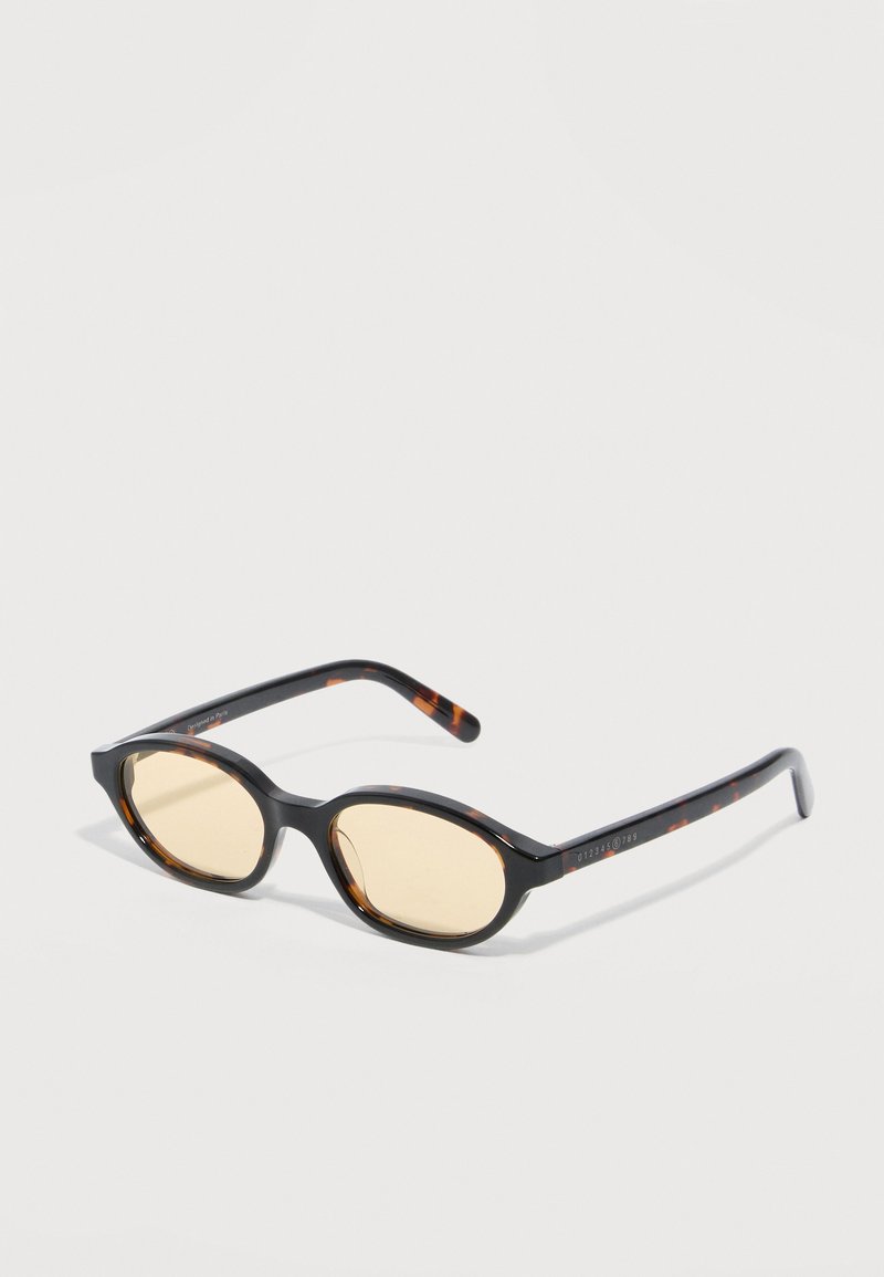 Black and tortoiseshell oval sunglasses with thin amber-tinted lenses and slim curved arms placed on a white surface.