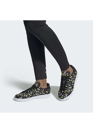 Ben i sorte leggings bærer sorte lavtop-sneakers med farverige blomsterprint og hvide såler på en lys grå baggrund.