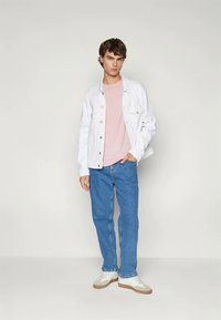 Mladý muž stojí, má na sebe bielu džínovú bundu, svetloružové tričko, modré džínsy a biele tenisky s hnedými podrážkami.