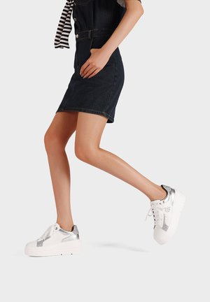 Sneakers bianchi con accenti argento, realizzati in materiale sintetico, caratterizzati da una suola spessa, lacci e lati texturizzati. Indossati con una gonna di denim scuro.