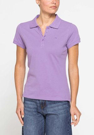 Donna che indossa una polo aderente color lavanda a maniche corte e jeans di denim blu su uno sfondo bianco semplice.