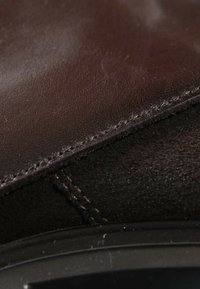 Gros plan d'une chaussure en cuir et daim marron foncé avec des coutures visibles et une partie de semelle noire.