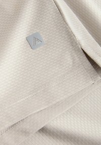 Gros plan sur un tissu beige texturé avec un petit patch logo carré réfléchissant comportant un motif triangulaire.