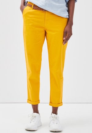 Personne portant un pantalon jaune vif à revers, une ceinture tressée marron, une chemise bleu clair et des baskets blanches, debout devant un fond uni.